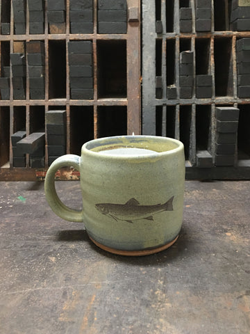 A light green ceramic mug with an image of a brook trout imprinted on the front. 