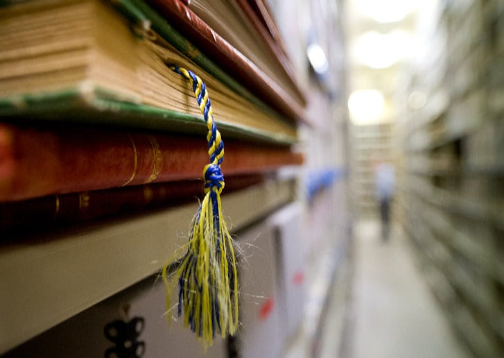 Focused photograph on five volumes on metal shelf. In the background, muted and unfocused, there is a long aisle of metal shelves with boxes.