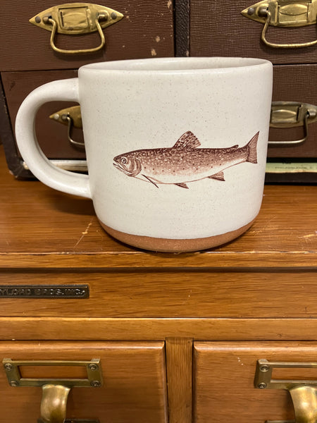 A cream-colored ceramic mug with an image of a brook trout imprinted on the front. 