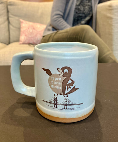 A light blue ceramic mug with Proud Robin logo imprinted on the front. Text on the Robin’s chest reads “It’s great to live in Michigan”