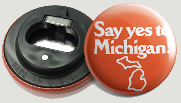 The front and back of a round bottle opener magnet featuring the logo for “Say yes to Michigan!” in bold white text with a white outline of Michigan underneath it on an orange background.