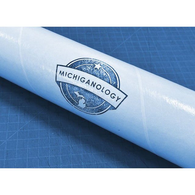 A Michiganology logo stamp on a white tube laying on top of a blue craft cutting mat.