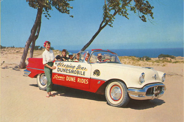 Front of a historical postcard showing a group of people in a red and white car on the top of a sand dune. The side of the car has text that reads “Sleeping Bear Dunesmobile Scenic Dune Rides”