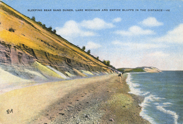 Front of a historical postcard showing the shoreline of Lake Michigan with a sand dune to the left. A woman bends down close to the water. Text on the top reads “Sleeping Bear Sand Dunes, Lake Michigan and Empire Bluffs in the Distance – 46”