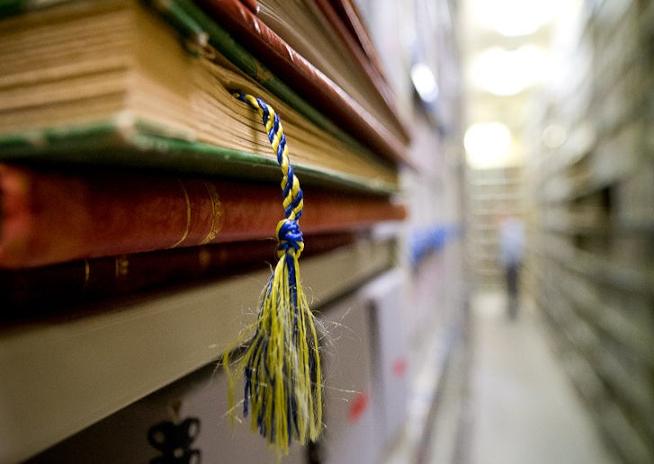 Focused photograph on five volumes on metal shelf. In the background, muted and unfocused, there is a long aisle of metal shelves with boxes.