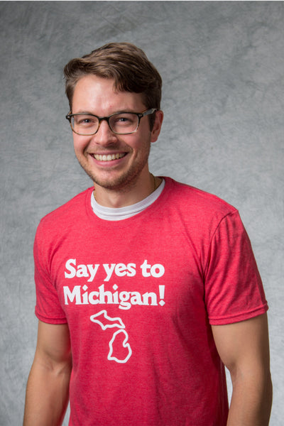 A smiling man wearing a red t-shirt with the words in white lettering “Say yes to Michigan!” with a white outline of the state of Michigan beneath it.