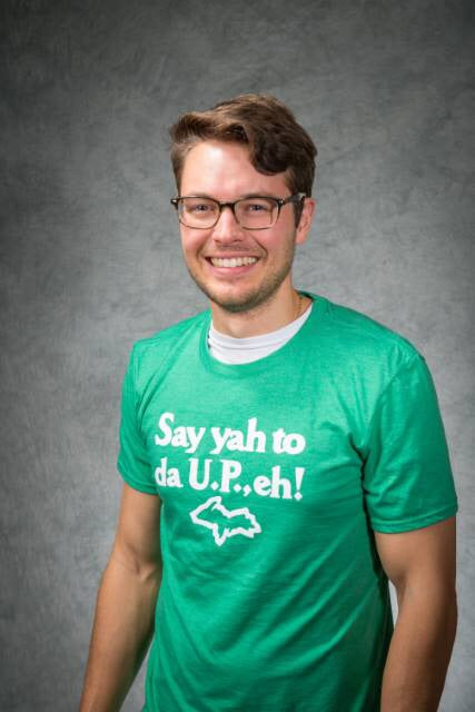 A smiling man wearing a green t-shirt with the words in white lettering “Say yah to da U.P., eh!” with a white outline of Michigan’s Upper Peninsula beneath it.