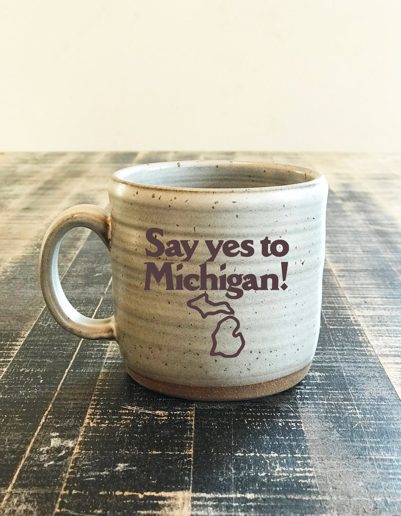 A cream-colored ceramic mug with the logo for Say yes to Michigan! imprinted on the front. 