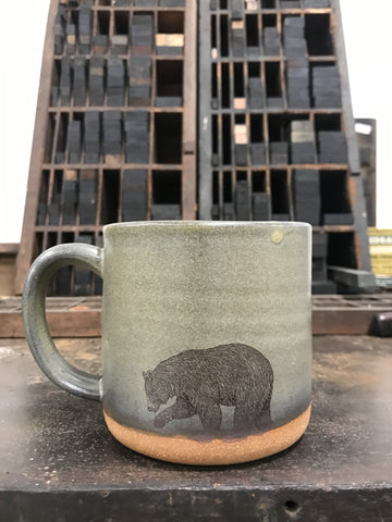 A moss green-colored ceramic mug with an image of a bear imprinted on the front.